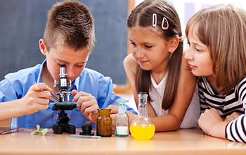 2 girls sitting by desk and a boy looking at a microscope
