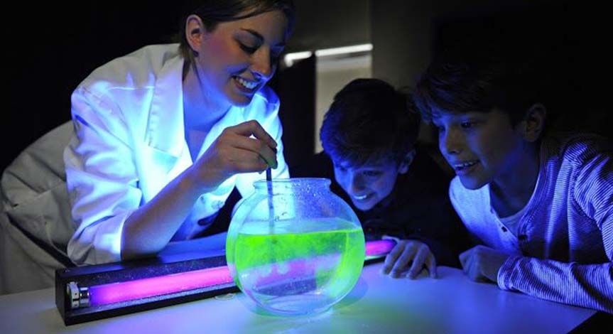 Mad Scientist pouring liquid that turns green with the use of a black light into a bowl of water with 2 kids looking on.