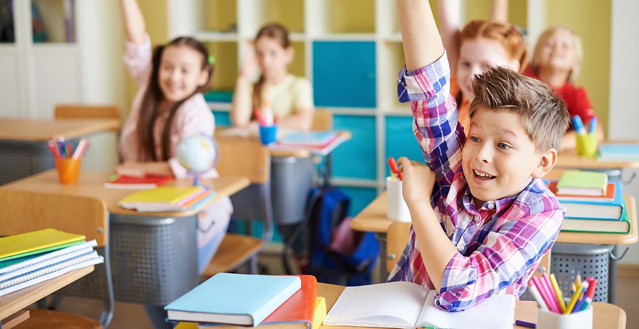 Boy in plaid shirt raising his hand in class with open notebook on his desk
